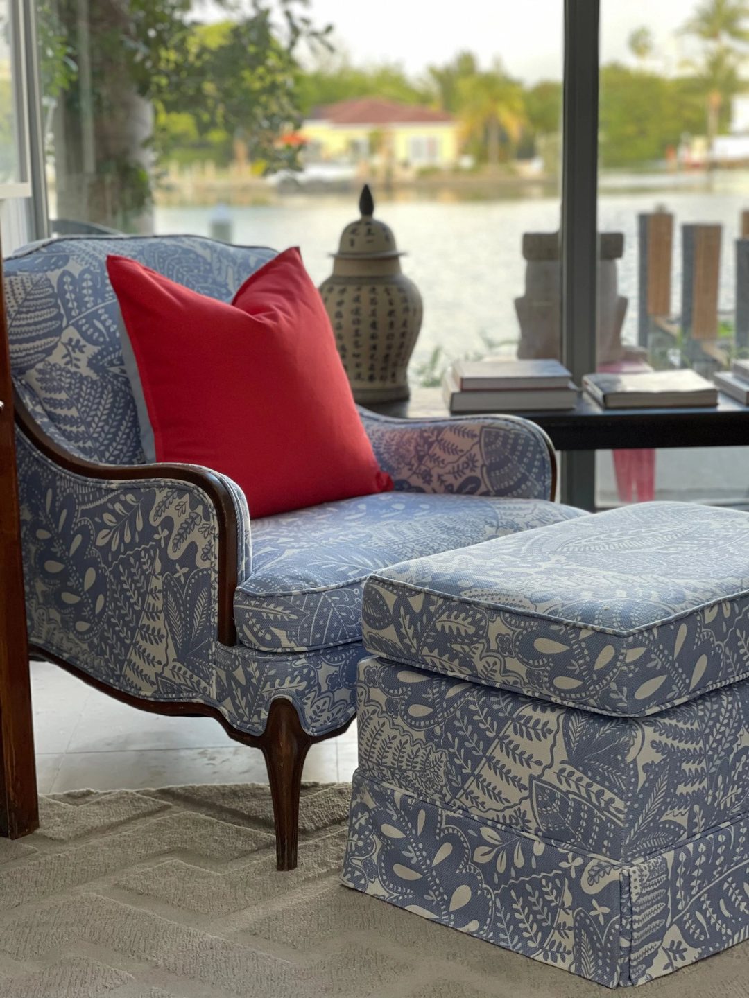 A wood-framed armchair and skirted ottoman in a blue-and-white botanical print, with a red pillow, in front of a window overlooking the water.