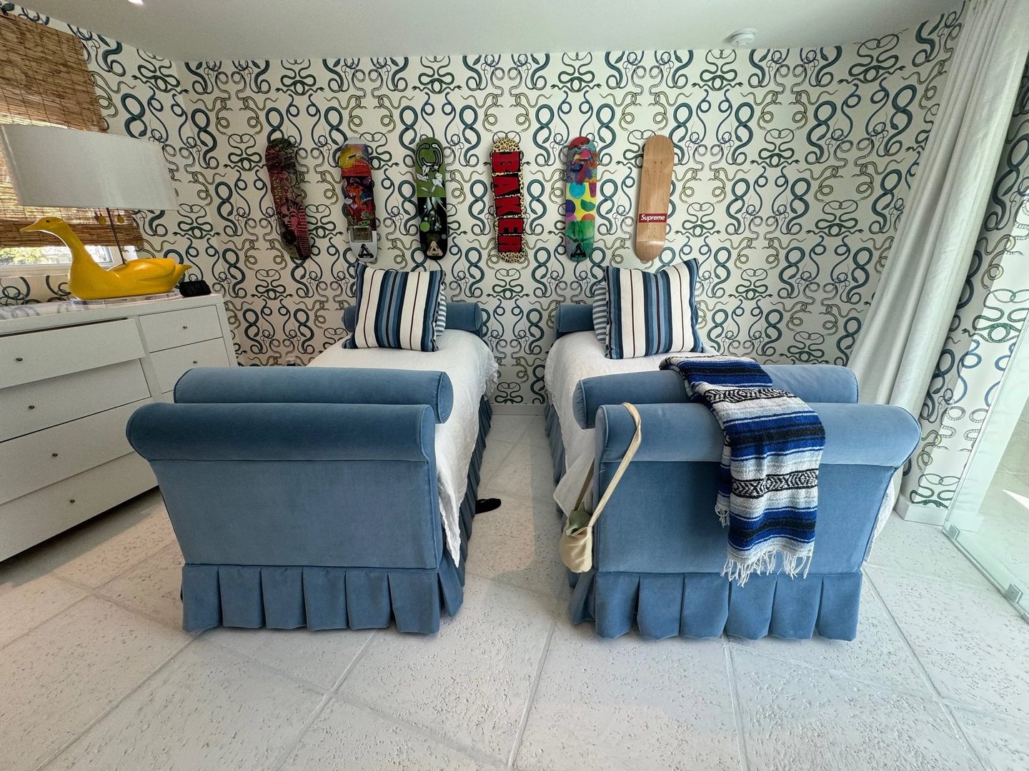 Two twin beds with rolled blue velvet frames and pleated skirts, with blue-and-white striped pillows, against green-and-white patterned wallpaper hung with skateboards.