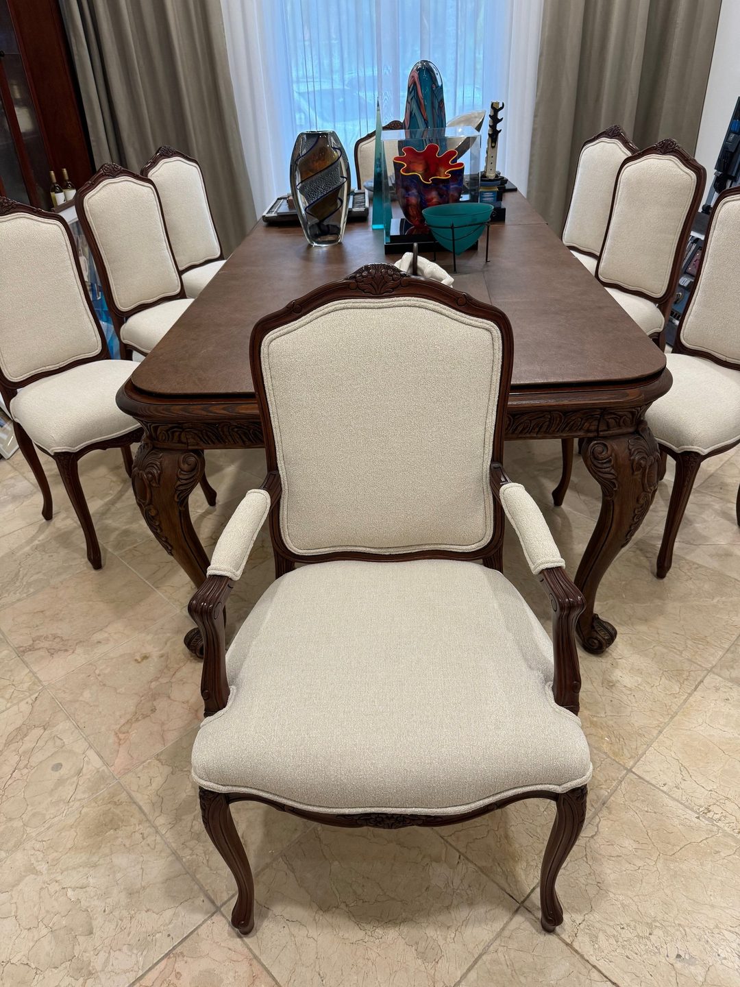 Carved wood dining chairs, including an armchair at the head, covered in cream fabric around a dark wood table.