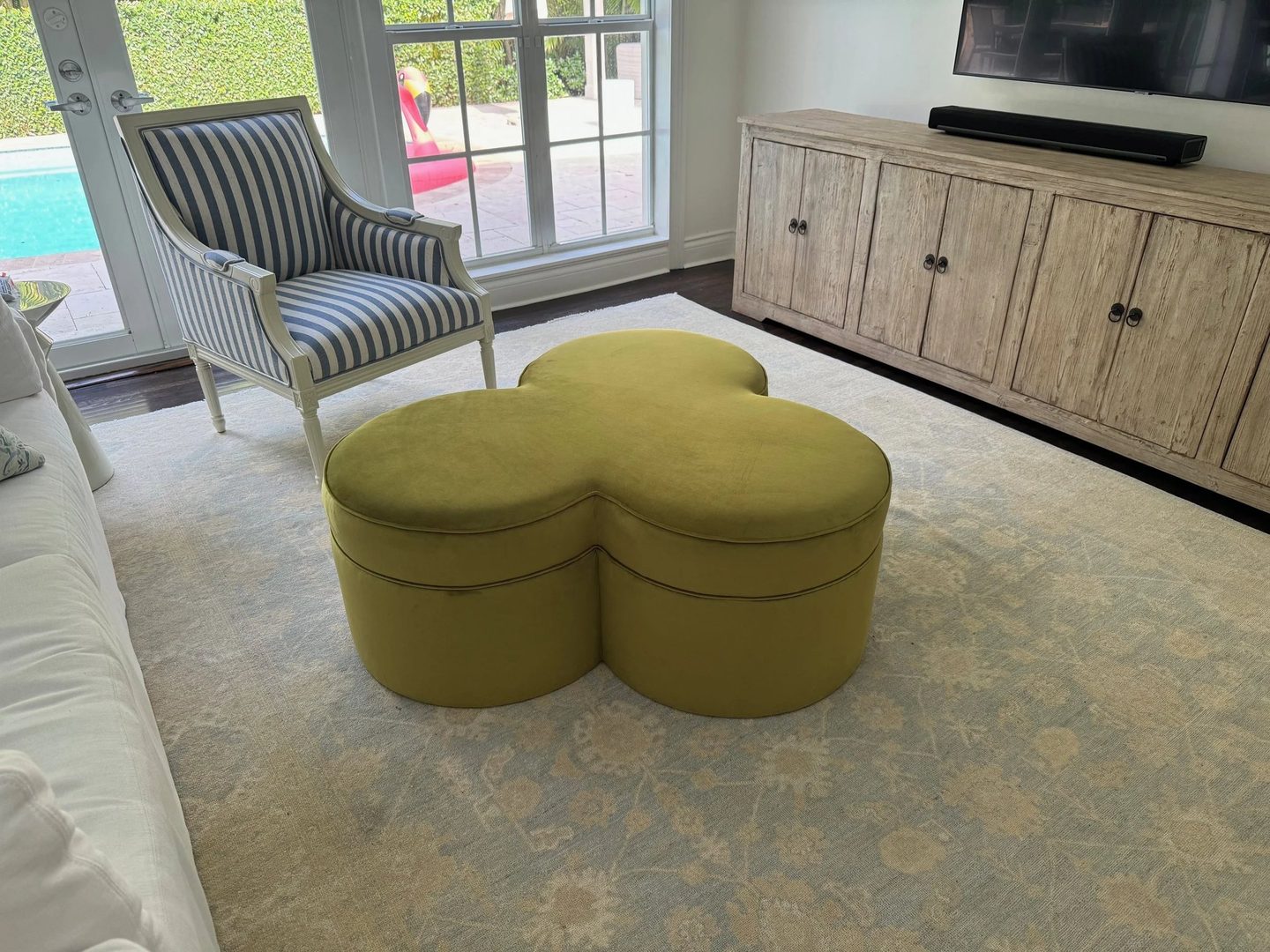 A clover-shaped ottoman in chartreuse velvet on a pale rug, with a blue-and-white striped armchair behind it.