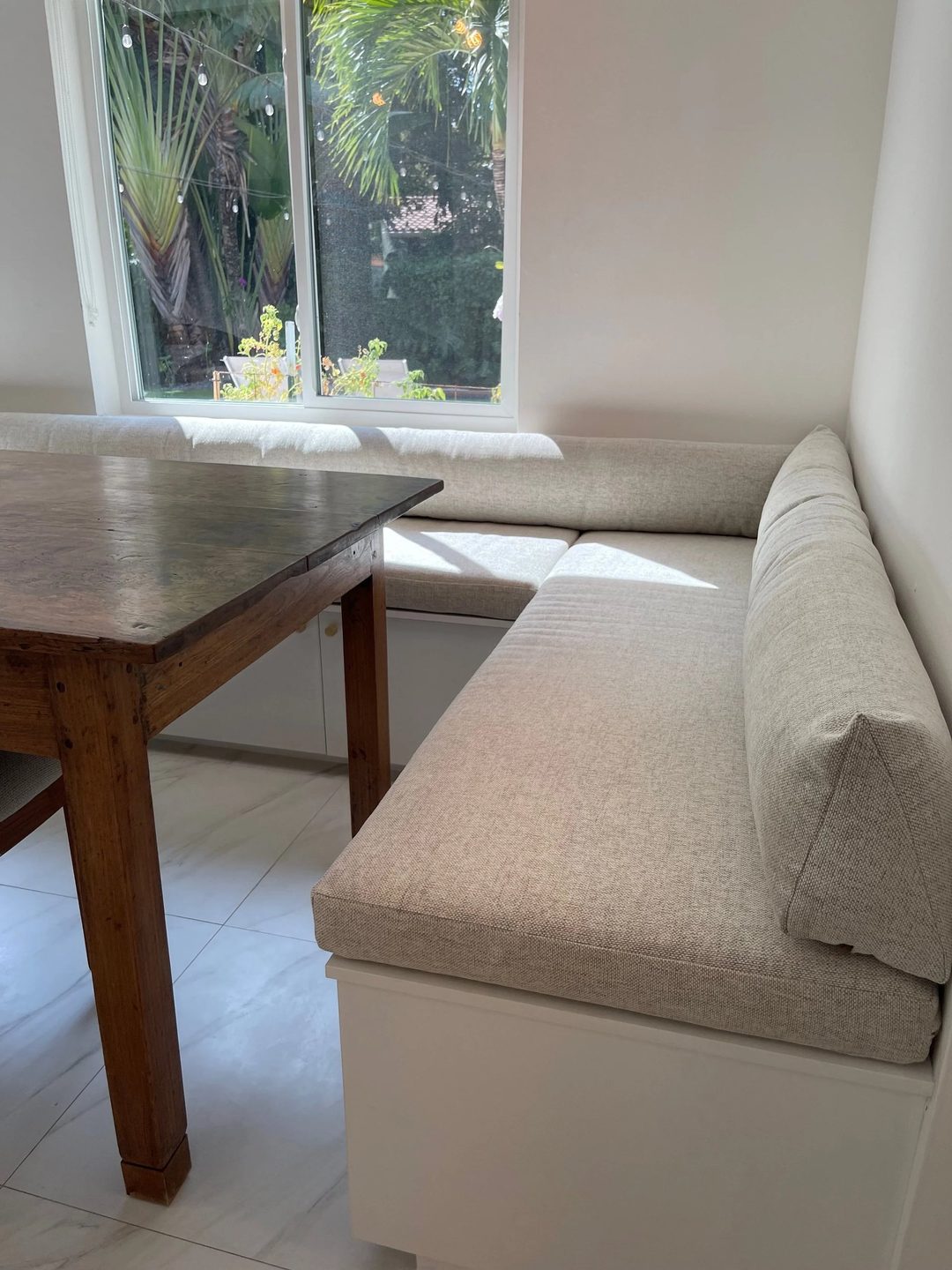 An L-shaped built-in banquette with oatmeal seat and back cushions on a white base, beside a wooden dining table and a window.