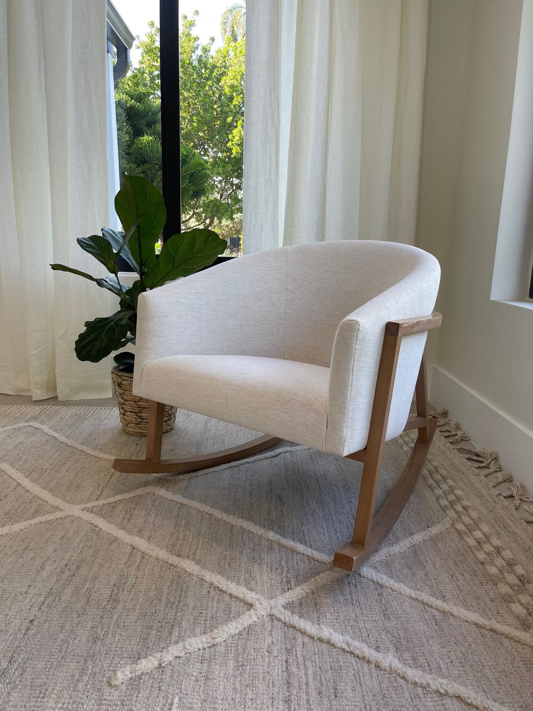 A cream upholstered rocking chair with a light wood frame, beside a fiddle-leaf fig and linen curtains.