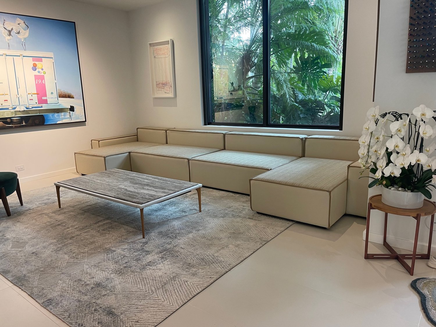 A long, low modular sectional in cream with brown piping, beneath a window of tropical plants, with a stone coffee table on a grey rug.