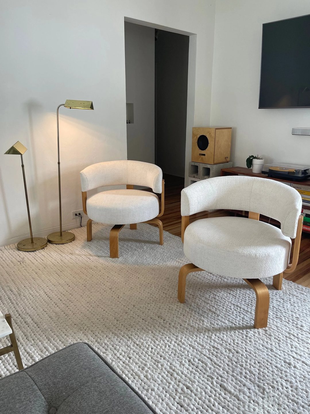 Two round-backed swivel chairs in textured cream fabric on bentwood bases, on a white wool rug beside two brass floor lamps.
