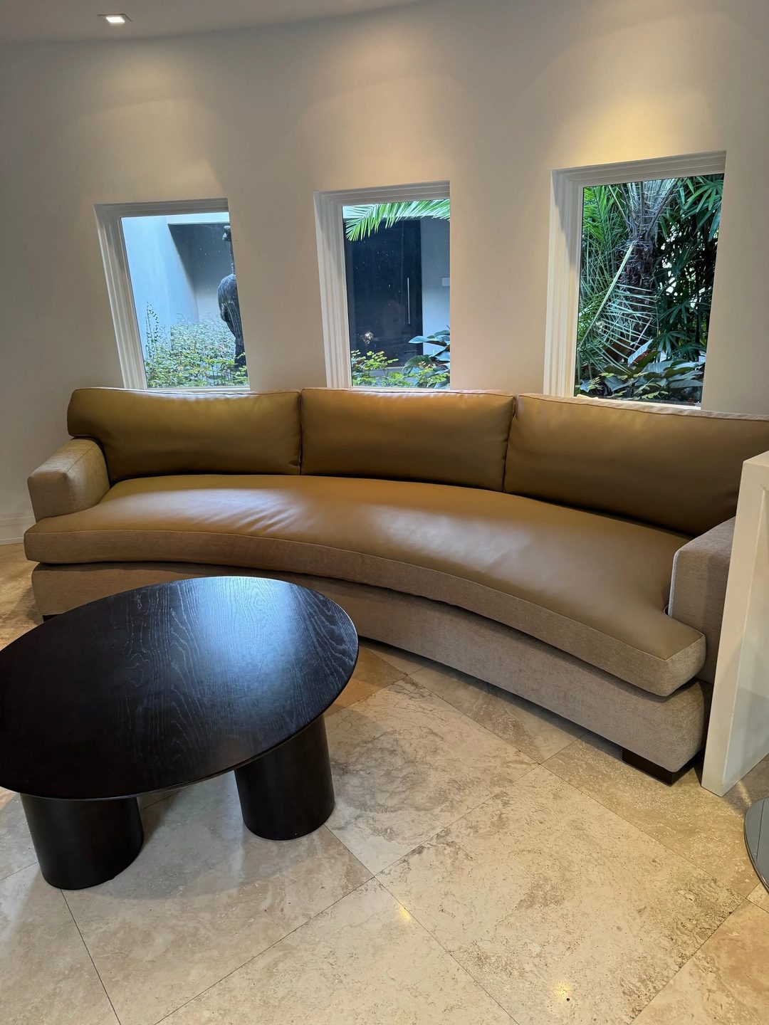 A long curved sofa with olive seat and back cushions on a sand-coloured base, in a round room with three windows.