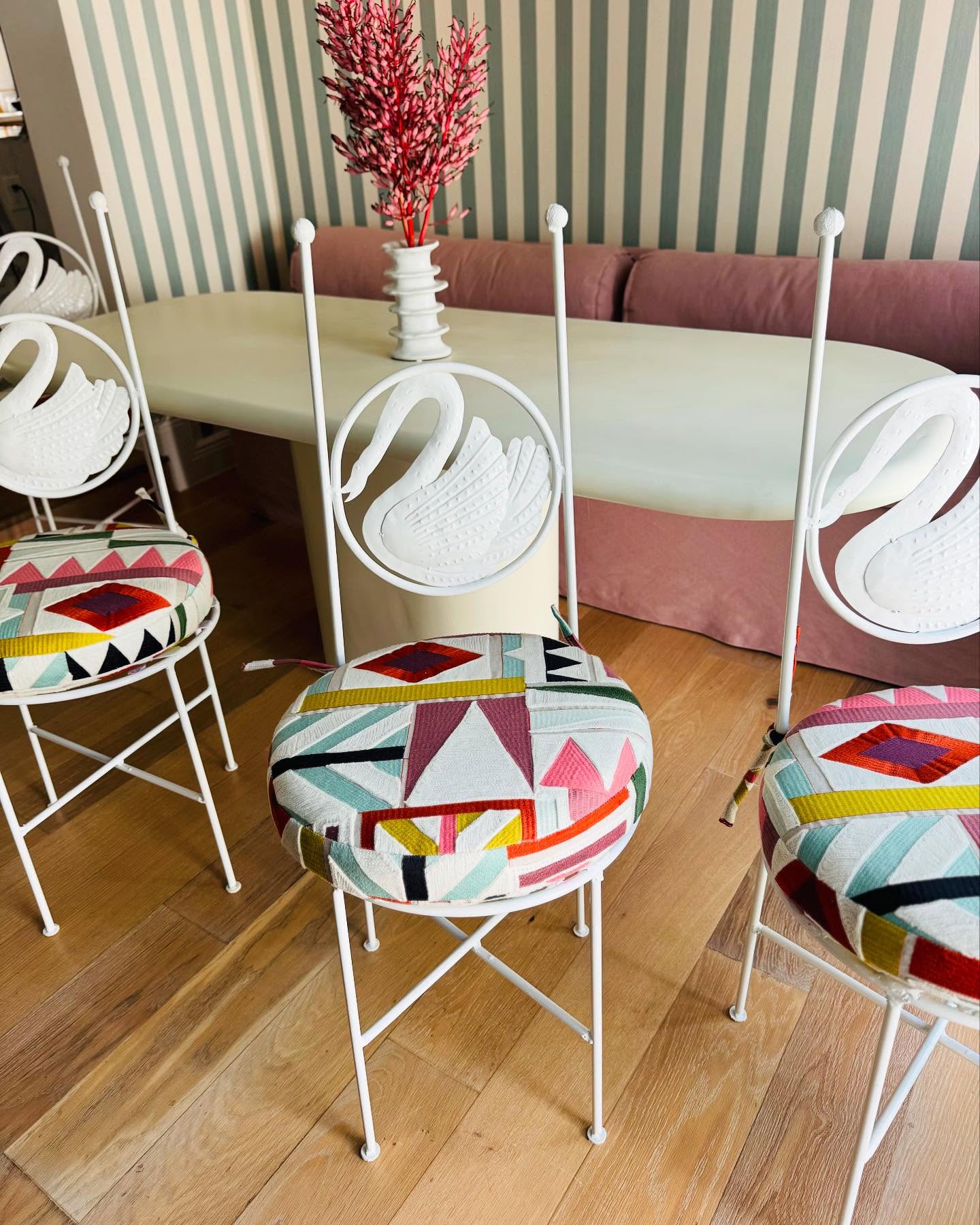 Four white swan-back chairs with bright geometric seat cushions around a pale dining table, with a dusty pink banquette behind them.