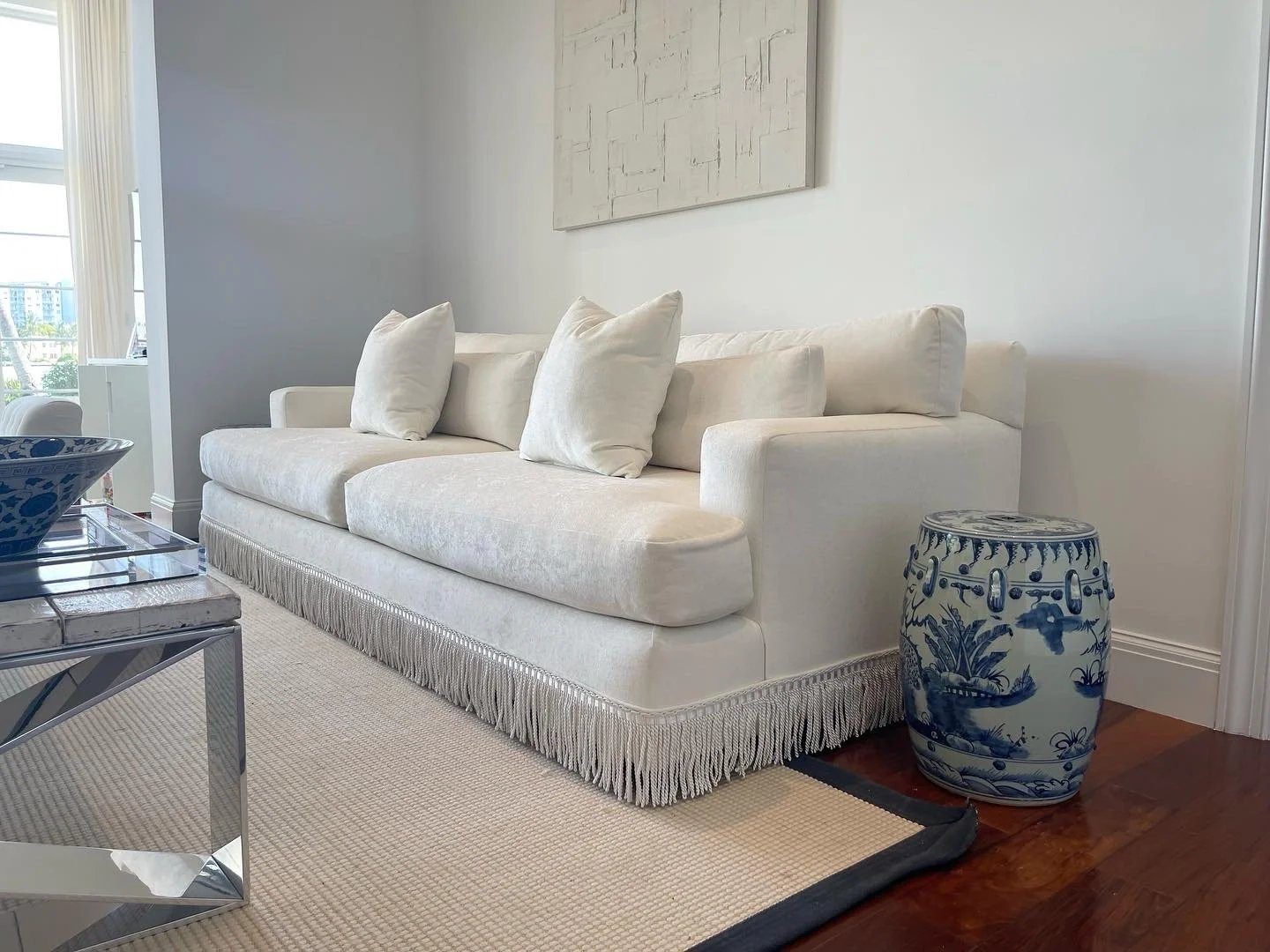 A white sofa with a long bullion fringe along its base, with a blue-and-white ceramic garden stool beside it.
