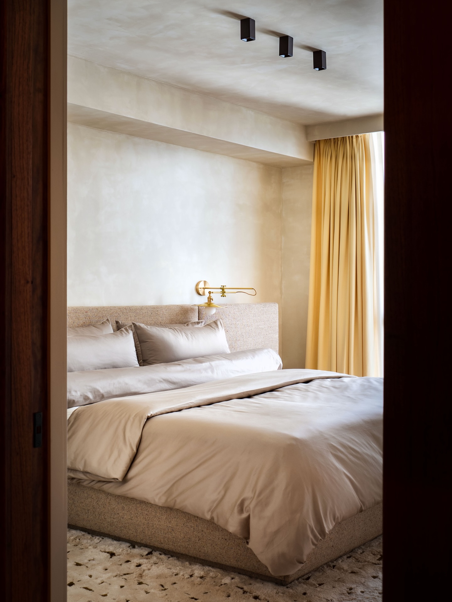 A bed with an upholstered oatmeal headboard and base, dressed in taupe satin linens, seen through a doorway, with yellow curtains beyond.