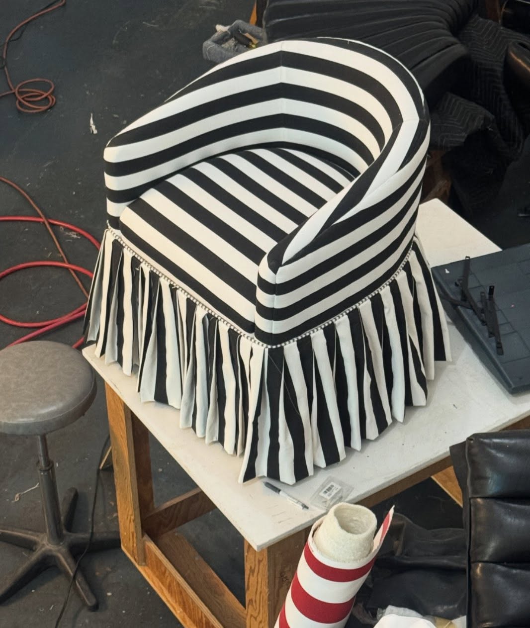 A small barrel-back chair in bold black-and-white stripes, with a pleated skirt and bead trim, sitting on a workshop table.