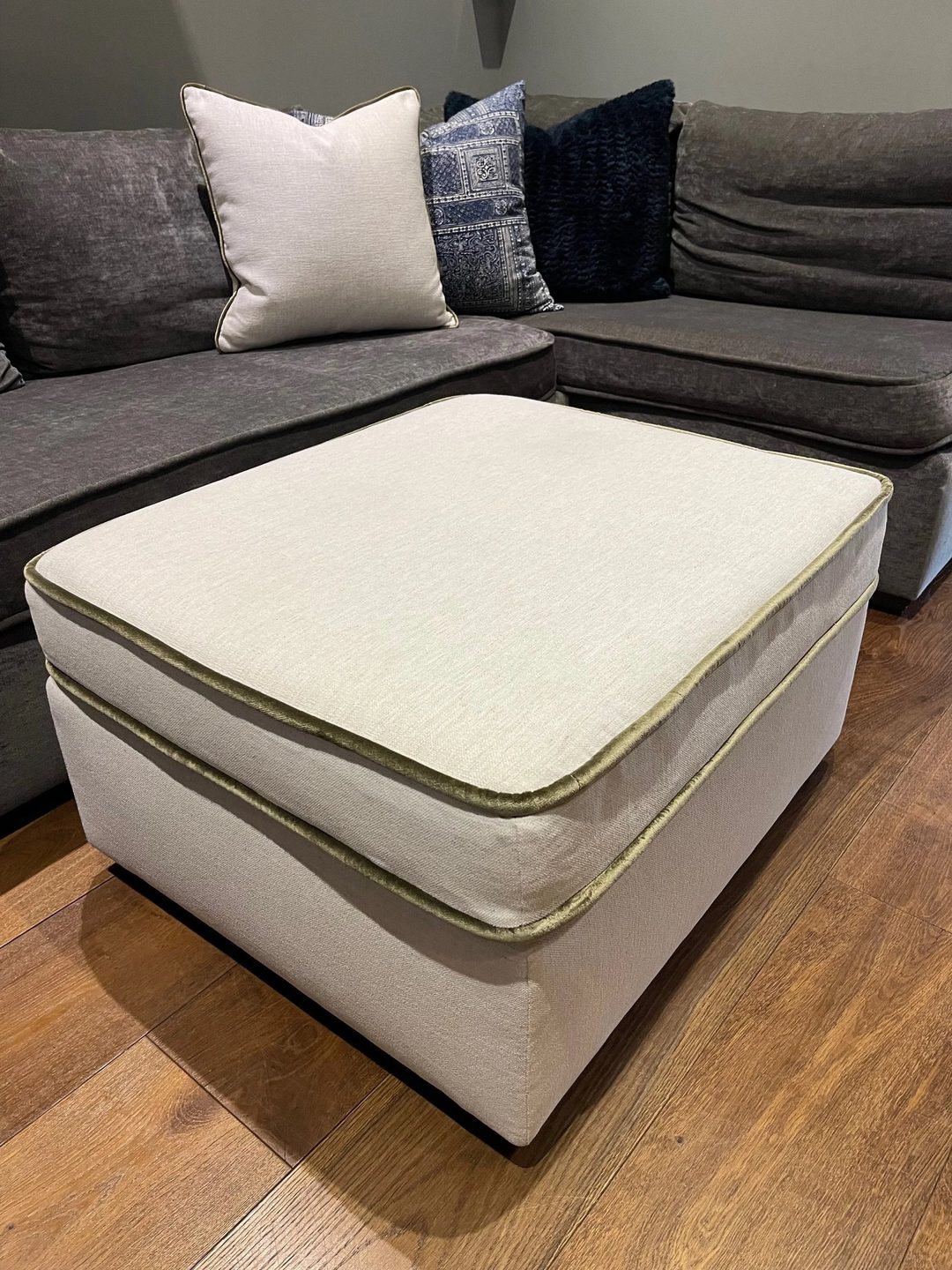 A square cream ottoman with olive velvet piping, in front of a charcoal sectional.