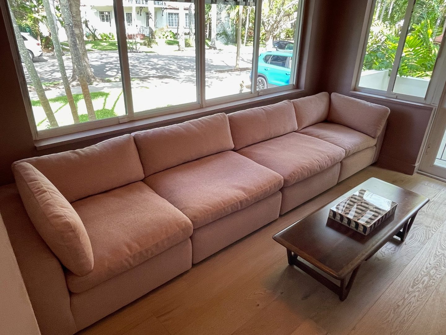 A long modular sofa in dusty rose fabric beneath a wide window, with a wood coffee table.
