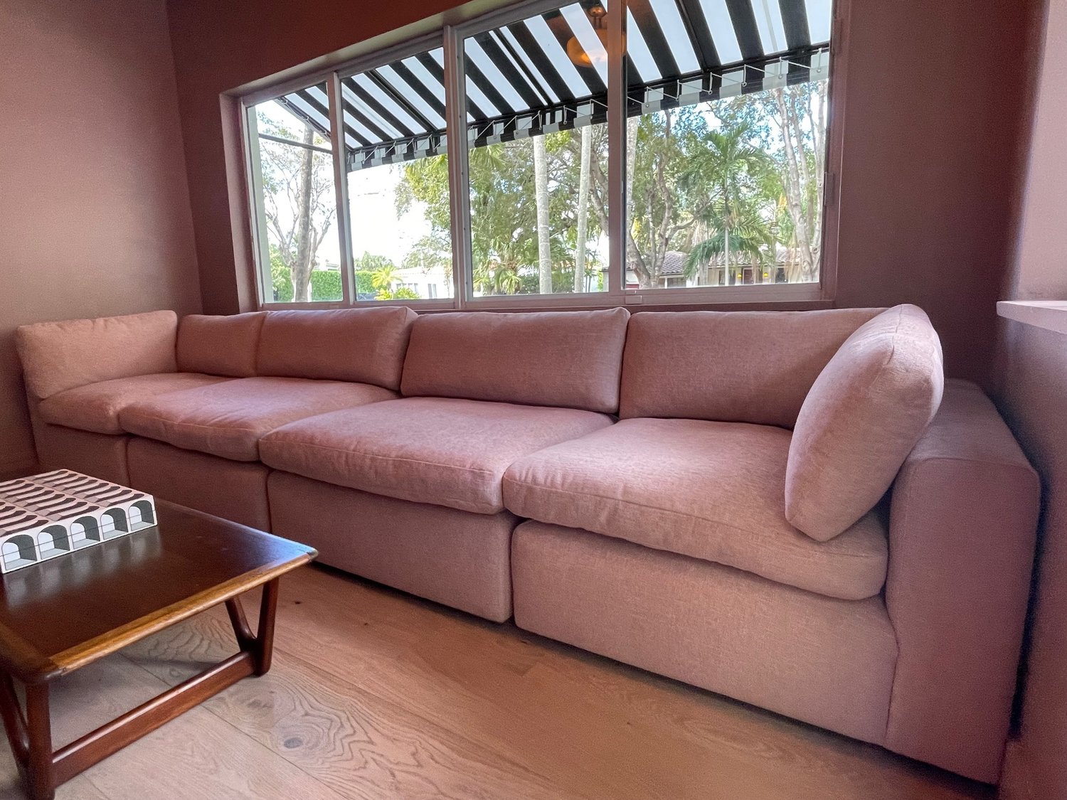 A long dusty rose sectional sofa beneath a wide window with a black-and-white striped awning outside.