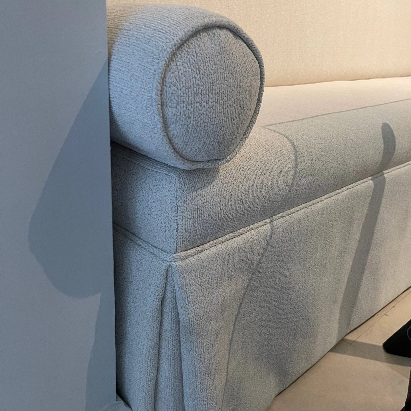 Close-up of a skirted bench in pale textured fabric, showing a round bolster, piped edges, and a kick pleat at the corner.