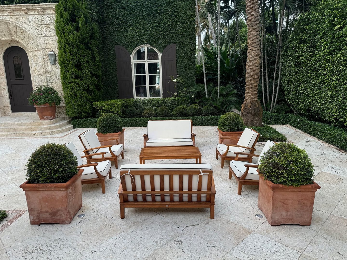 Teak outdoor sofas and armchairs with white cushions around a coffee table in a stone courtyard, with potted topiaries.