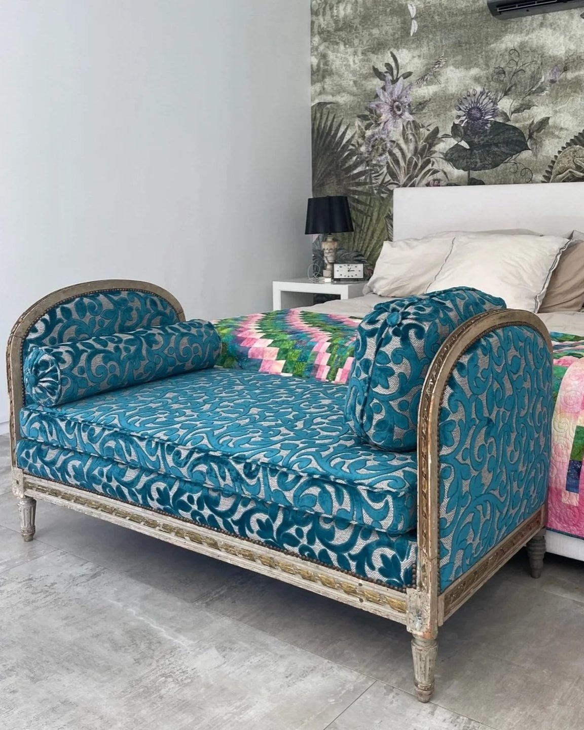 A painted wood daybed upholstered in teal and silver cut velvet, with a matching bolster and pillow, at the foot of a bed with a patchwork quilt.