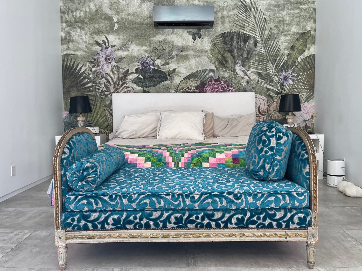 A carved, painted daybed upholstered in teal cut velvet with a bolster and a round pillow, in front of a bed and a botanical mural.