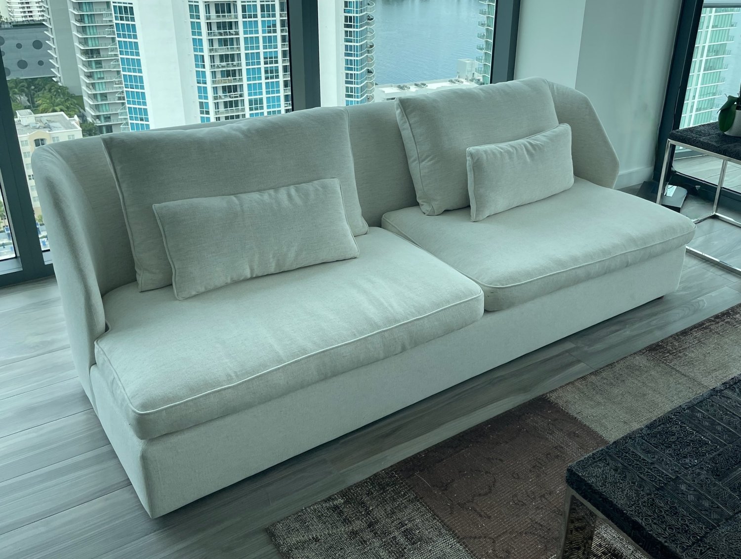 A low white sofa with loose back cushions and lumbar pillows, in front of floor-to-ceiling windows overlooking the water.