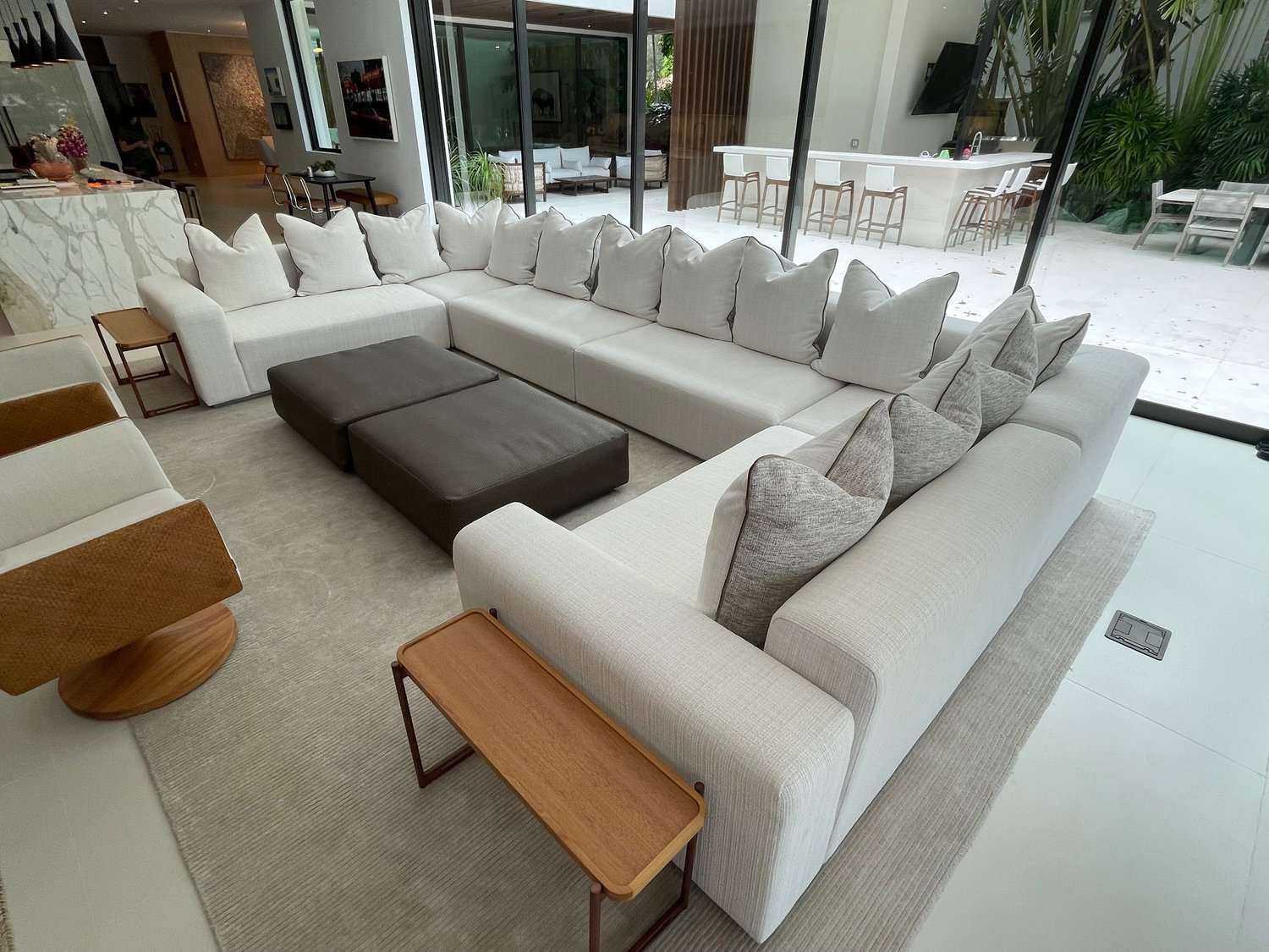 A large U-shaped white sectional with grey and white pillows around two charcoal ottomans, open to a pool terrace.
