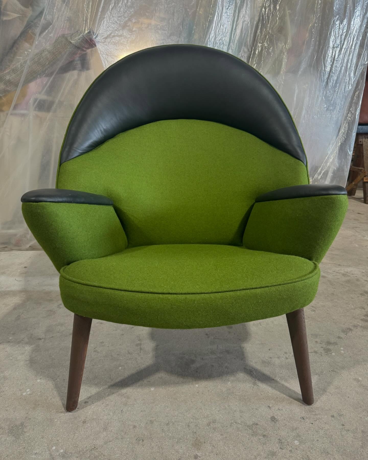 A restored womb chair in green fabric with black leather on the crest and arms, and tapered wood legs.