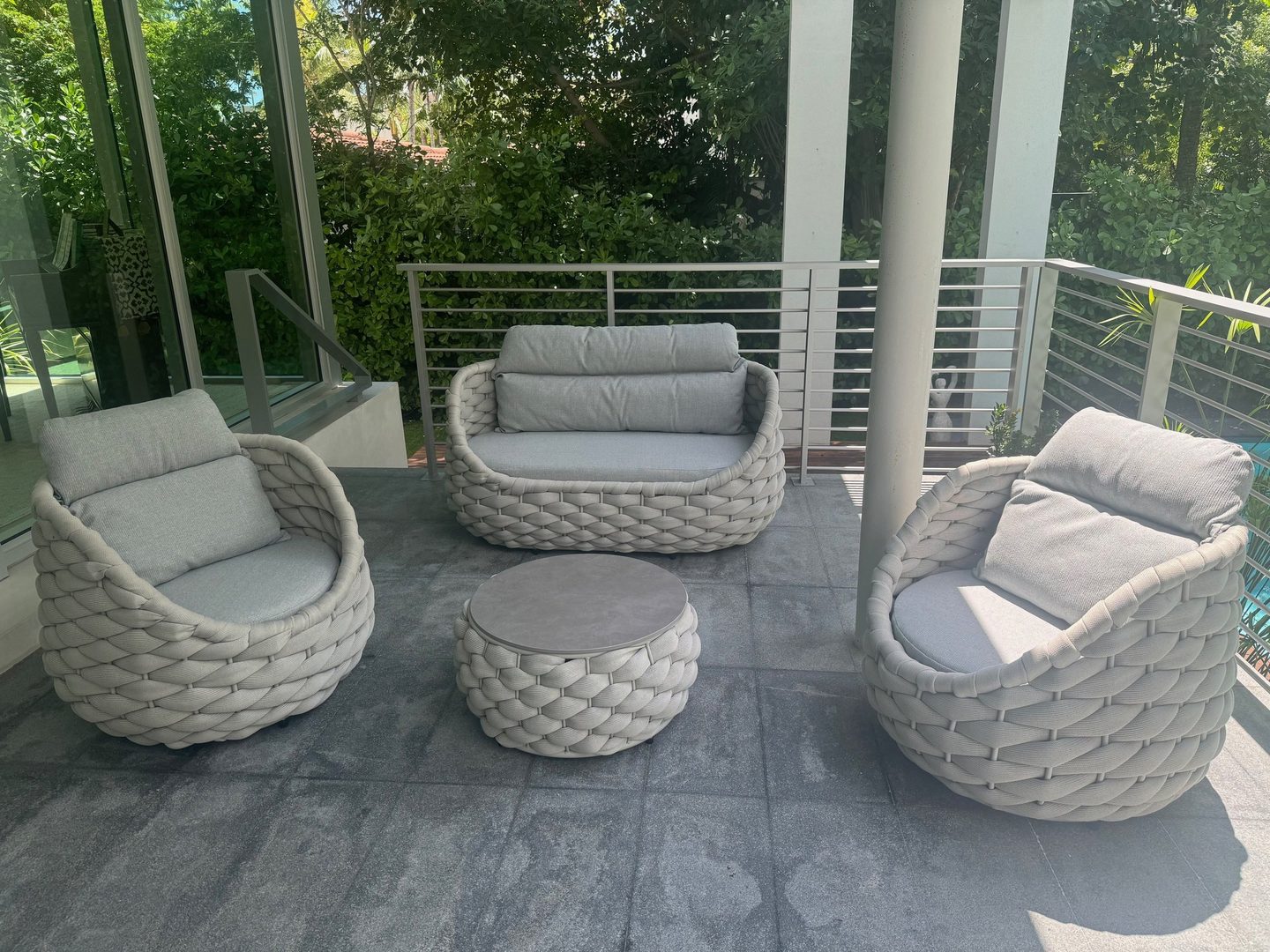 A woven outdoor loveseat, two lounge chairs, and a drum table with soft grey cushions, on a shaded terrace.